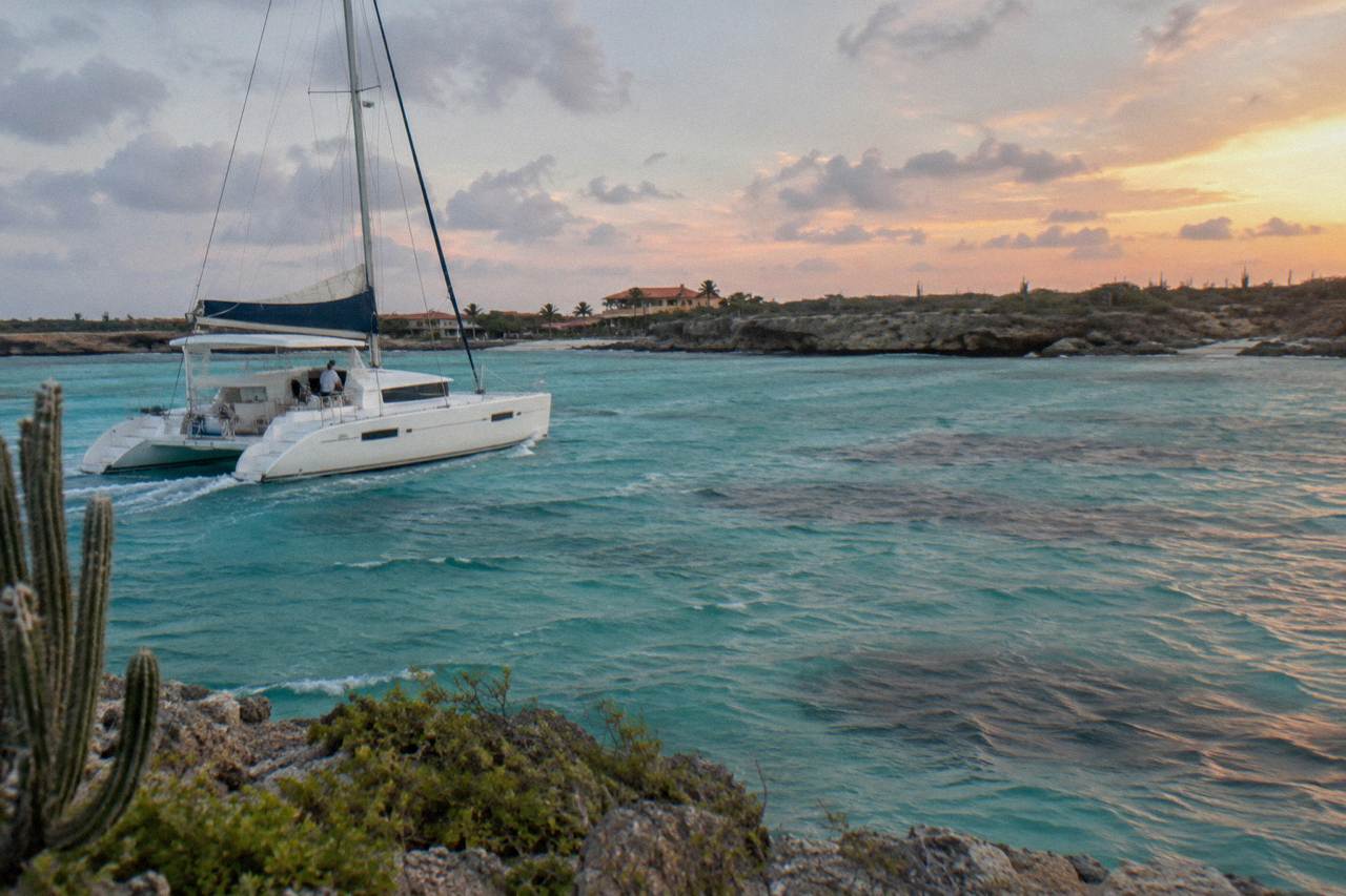 Catamaran sails across calm water under an orange and pink evening sky near the Aruba coast