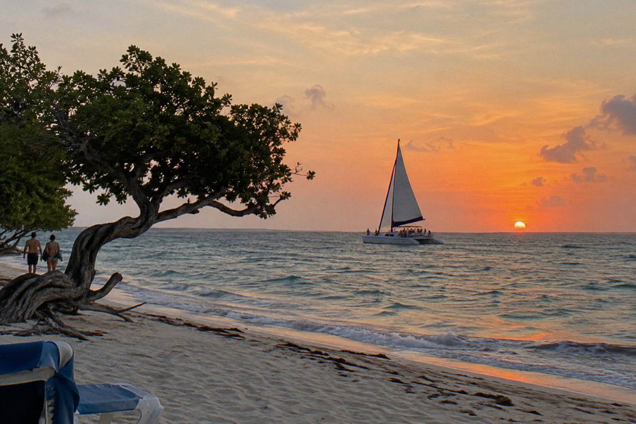 A catamaran sail on calm water under a wide orange and pink evening sky along an island coastline