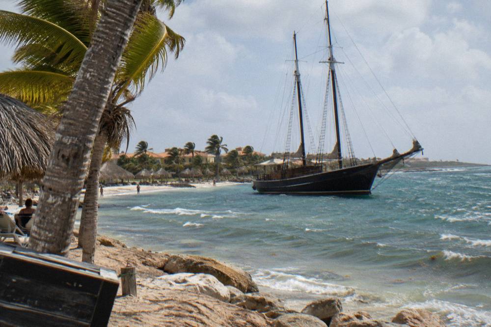 Wooden pirate style schooner with tall masts moored beside a palm lined beach in late afternoon light