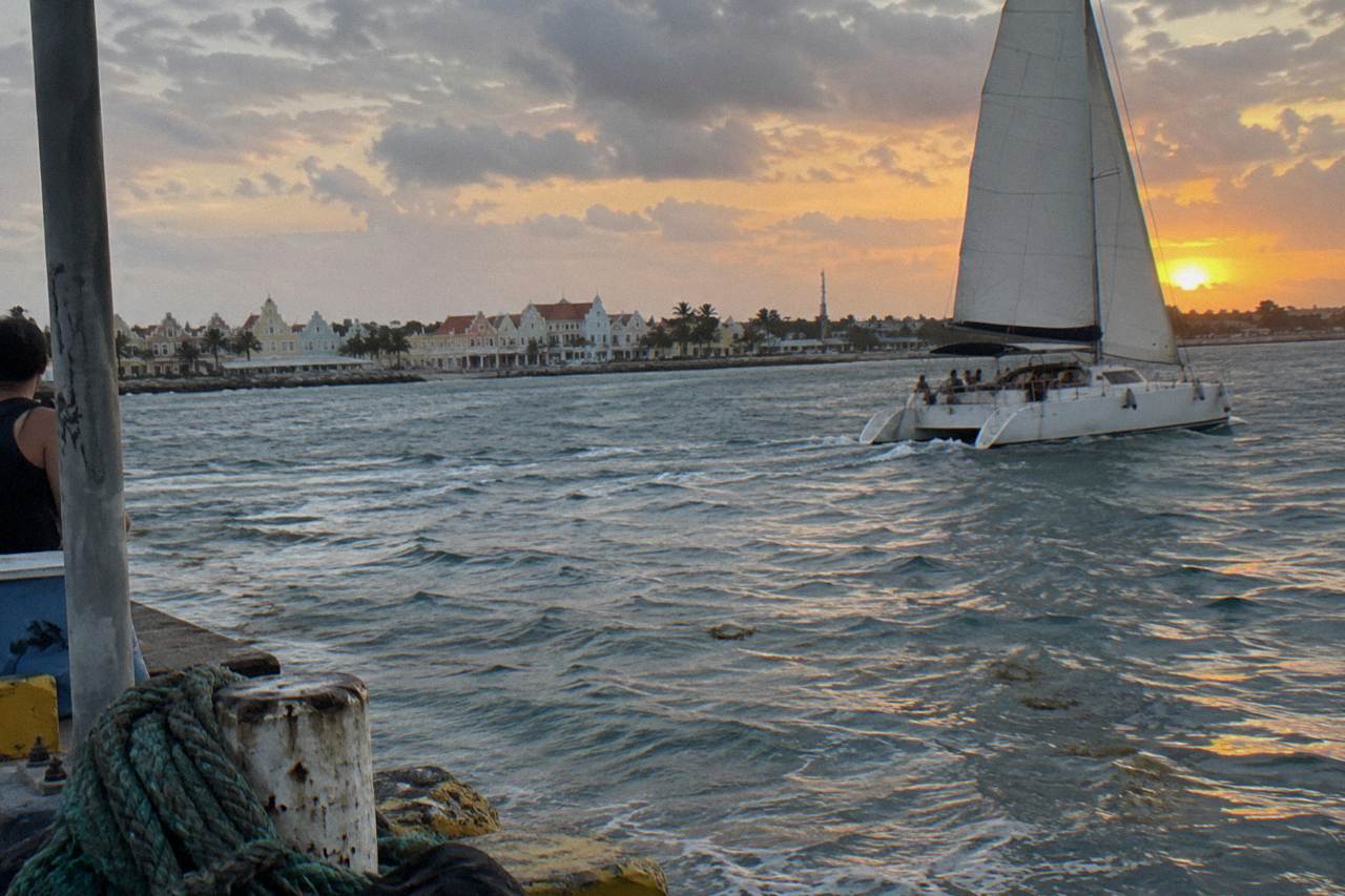 Catamaran under sail off the Aruba coast at dusk with an orange sky over calm water