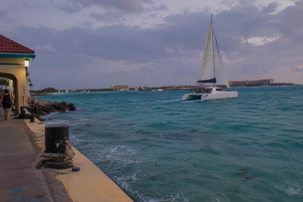 A wide catamaran under sail off a Caribbean coast with calm turquoise water and a dimming evening sky