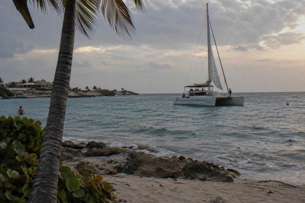 A catamaran with sails partly raised drifting on nearly flat water at dusk