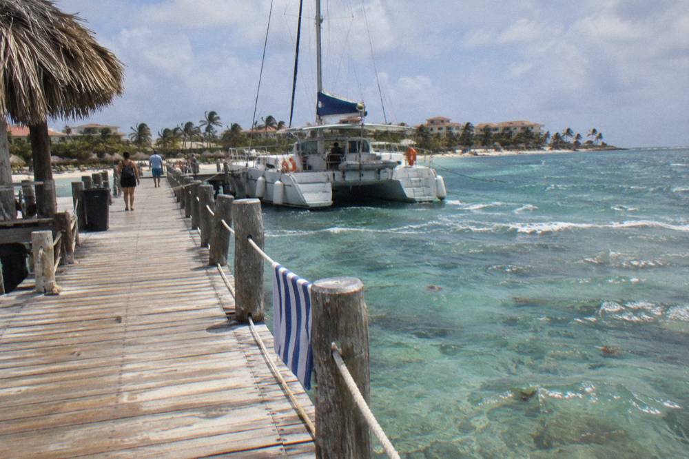 Wooden pier extending over clear shallow water toward a moored catamaran under a bright sky