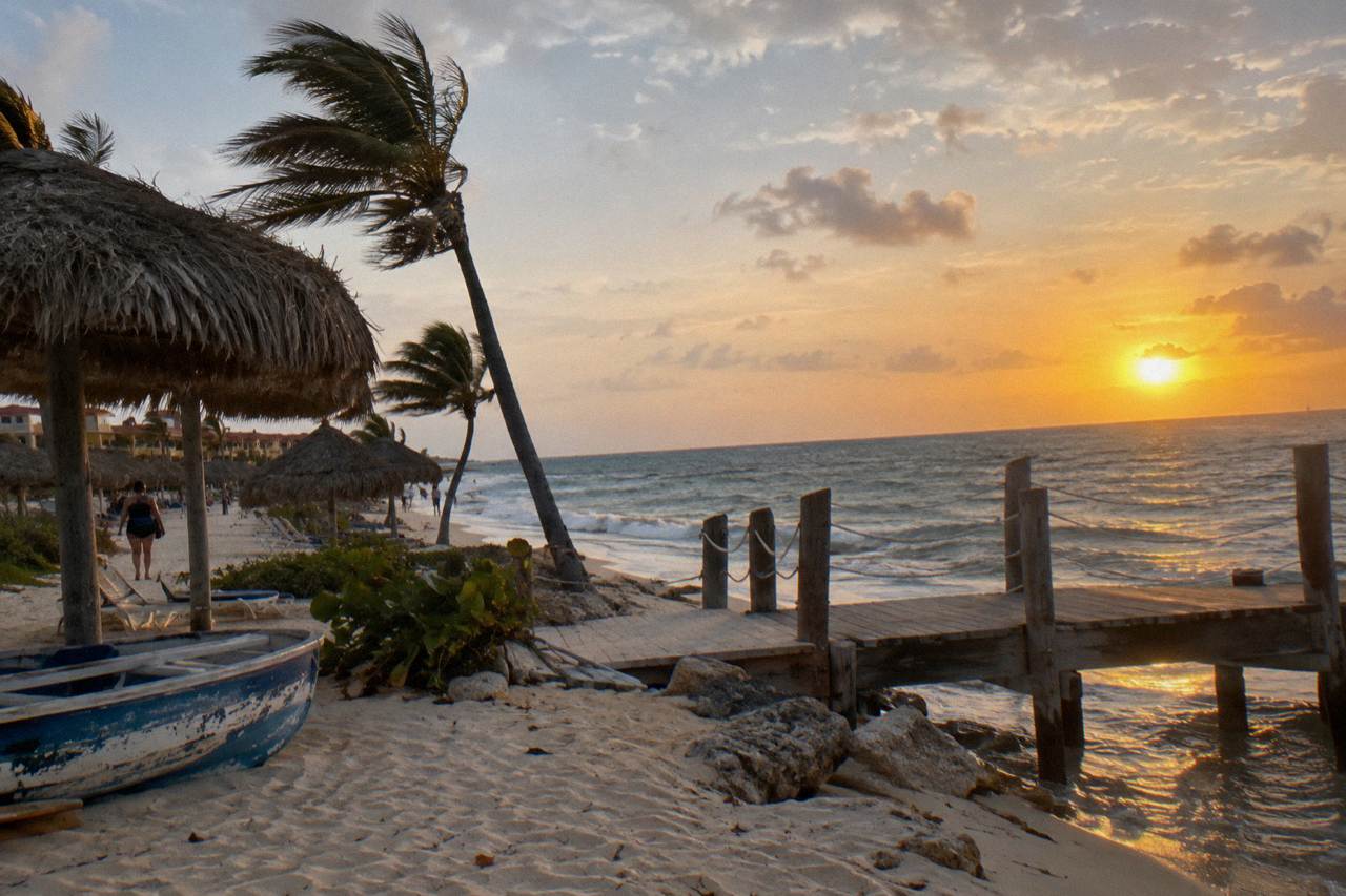 Golden sunset over calm Caribbean water beside a wooden beach pier with thatched shade structures