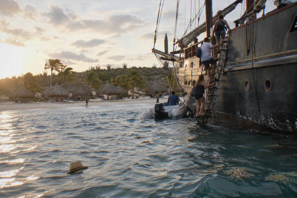 Pirate schooner anchored near a beach with passengers boarding by ladder at golden hour