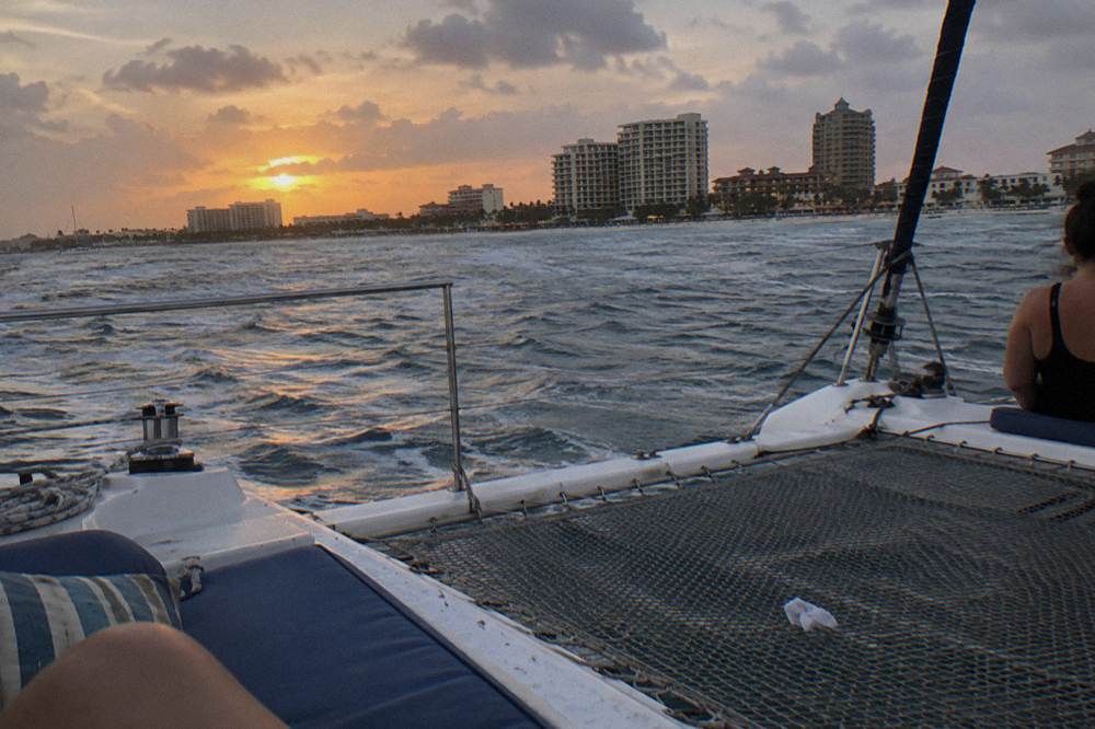 Open deck of a catamaran at dusk with the coastline glowing under an orange and purple sky