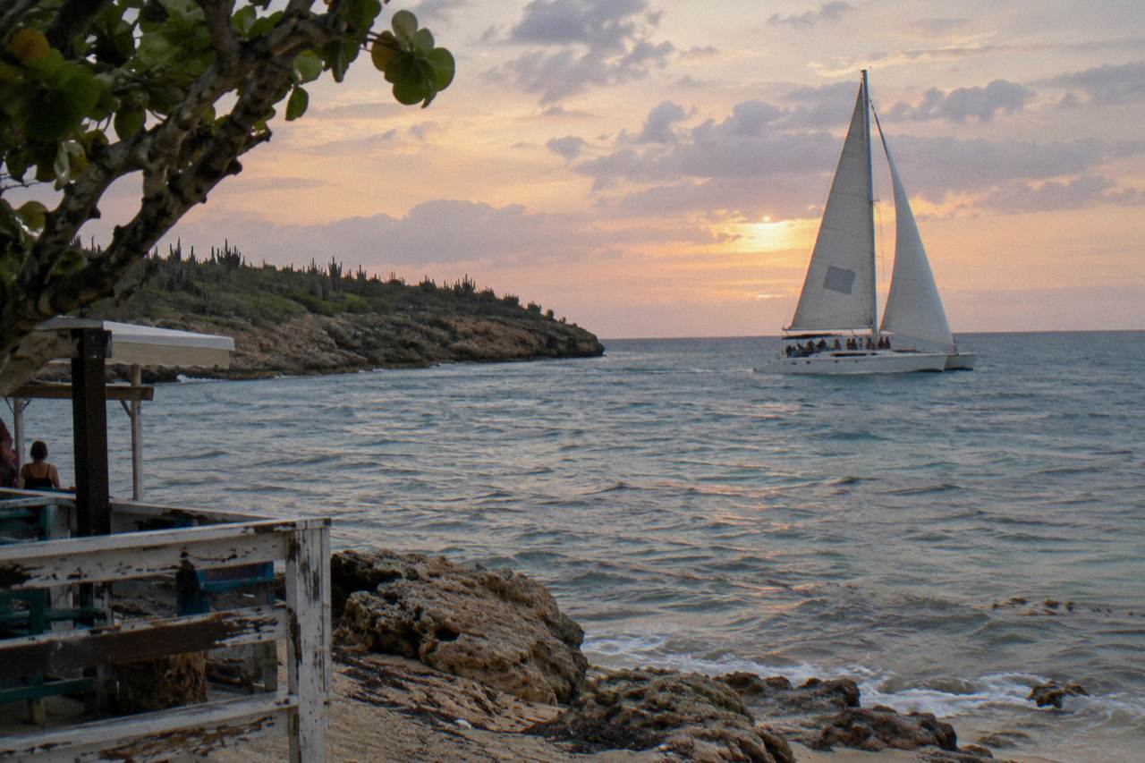 Catamaran sails across calm water under an orange and pink evening sky