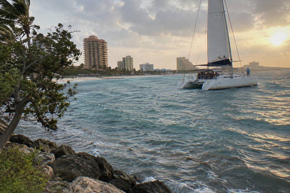 A catamaran under sail along the Palm Beach coast as the sun drops toward the horizon.