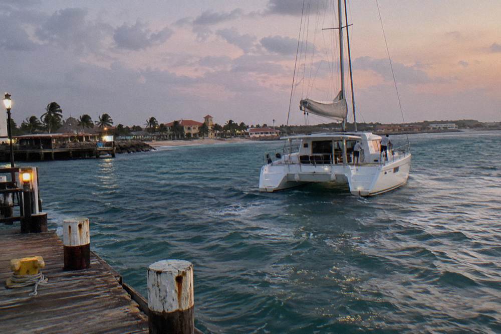 Catamaran under sail returning to a wooden pier under a fading pink and violet evening sky