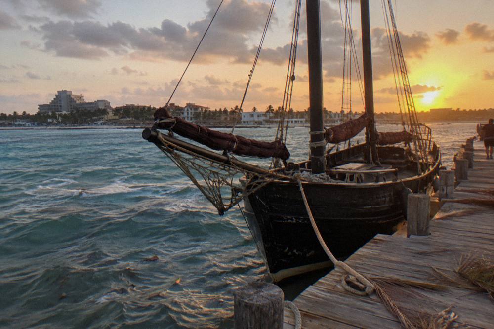 Wooden pirate-style schooner with masts against an orange evening sky at a beach pier