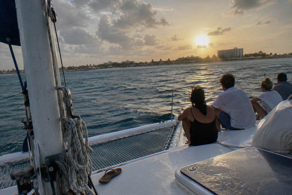 A wide catamaran deck with a small group seated together as the sun lowers over calm Caribbean water