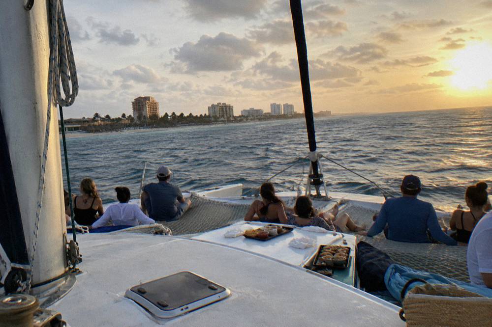 A trimaran heeling slightly with guests seated along its nets and the sun low over open water