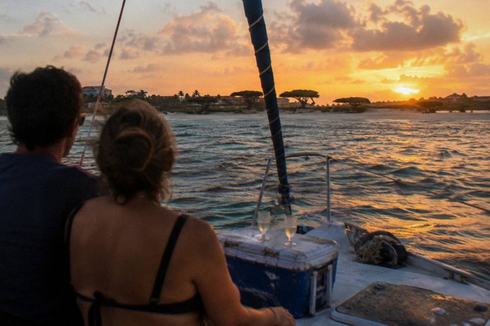 A couple seated at the bow of a small sailing boat with the sun setting behind them over calm water
