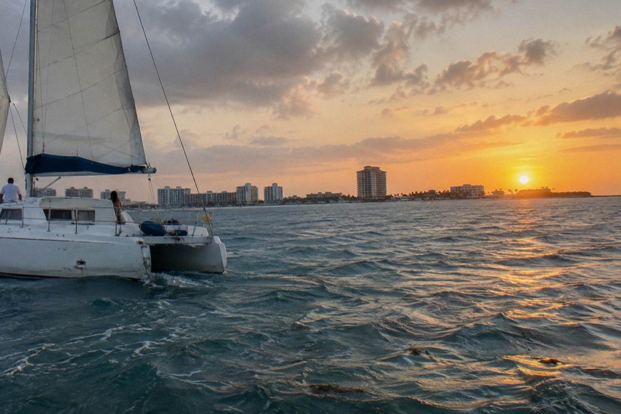 A white catamaran under sail on calm Caribbean water as the sun sets over the Aruba coastline