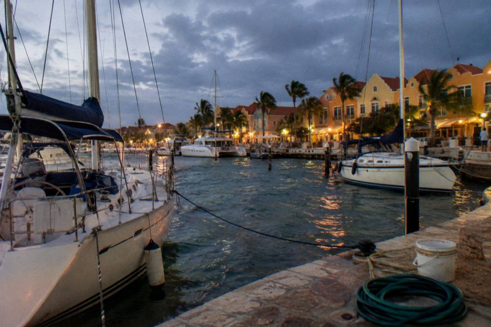 Boats anchored near a marina at dusk with dark blue sky and warm lights along the waterfront