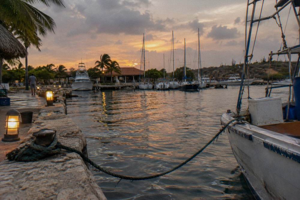 Small marina at dusk with masts and calm harbor water reflecting orange sky