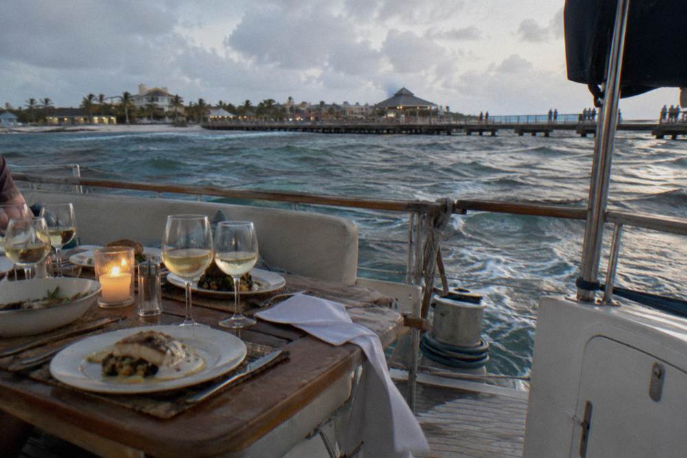 A dinner table set on a boat deck with glasses and candlelight near open water