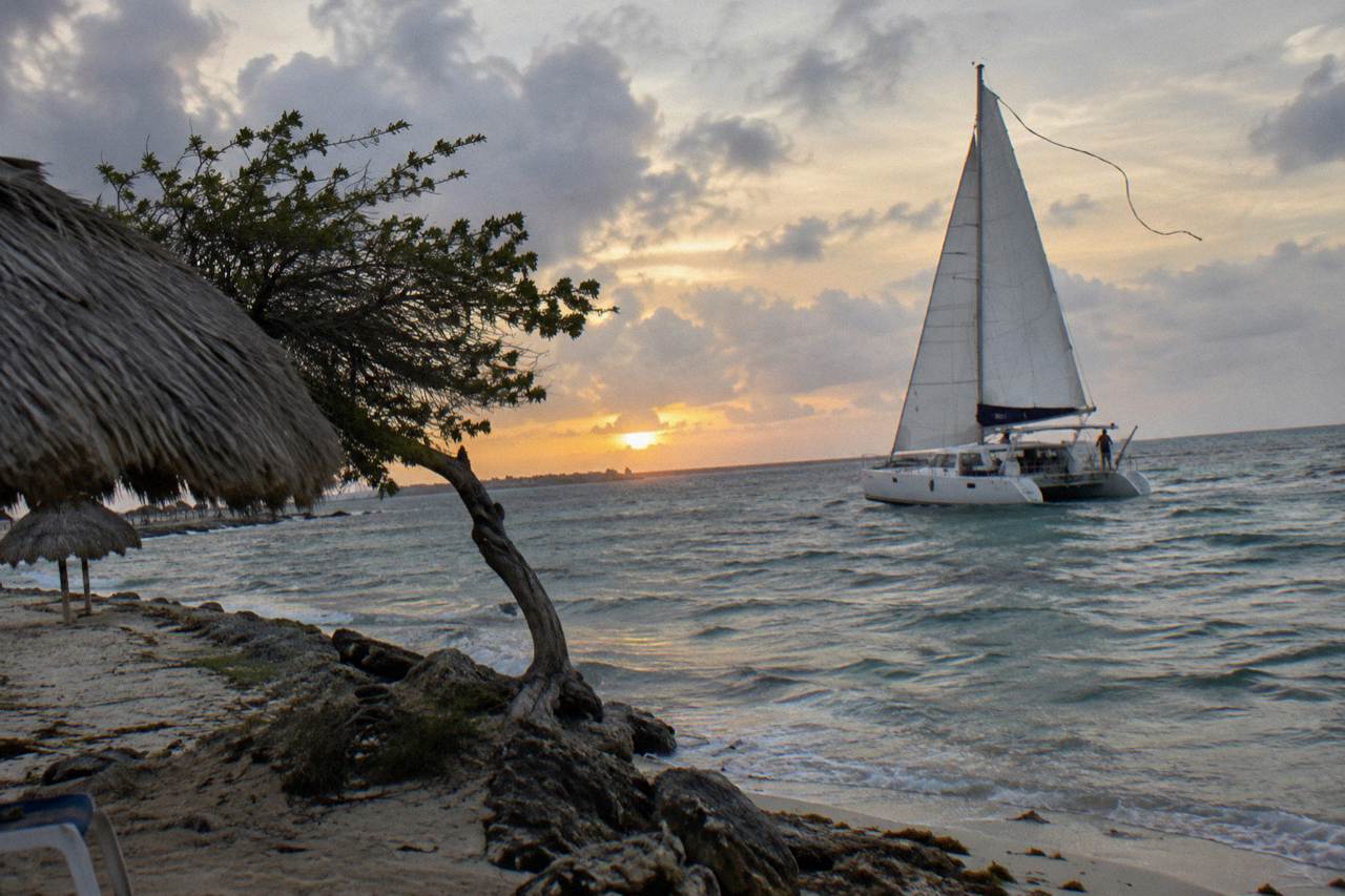 Catamaran under sail on calm Caribbean water as the sun sets over the Aruba coastline