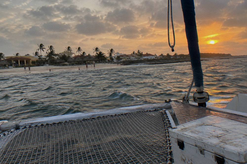 A wide catamaran deck with netting at the bow facing an orange evening sky
