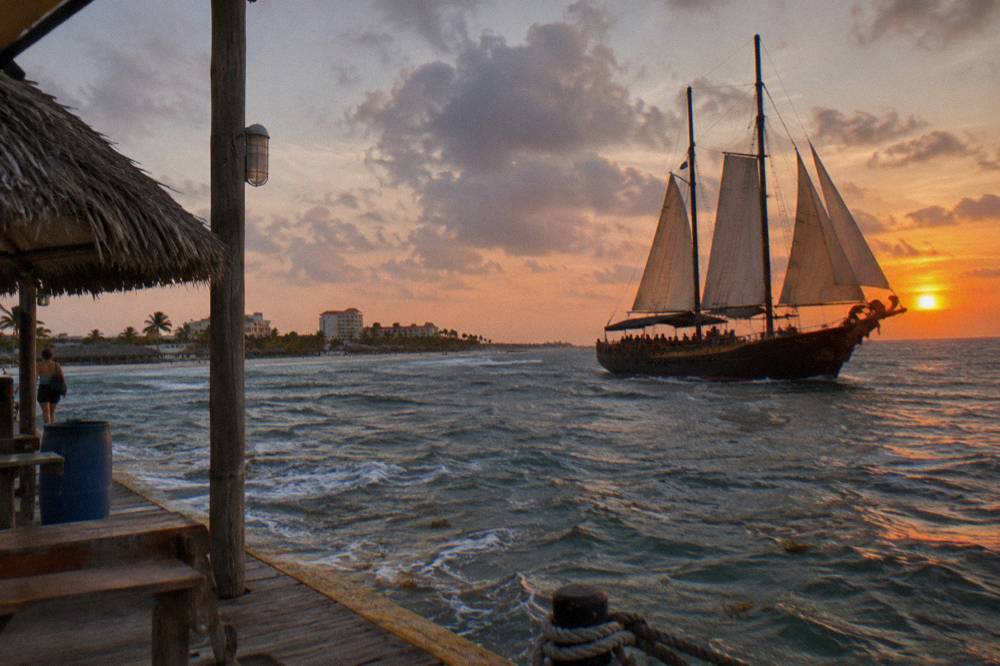 A wooden pirate schooner under sail with rigging and a figurehead silhouette at sunset