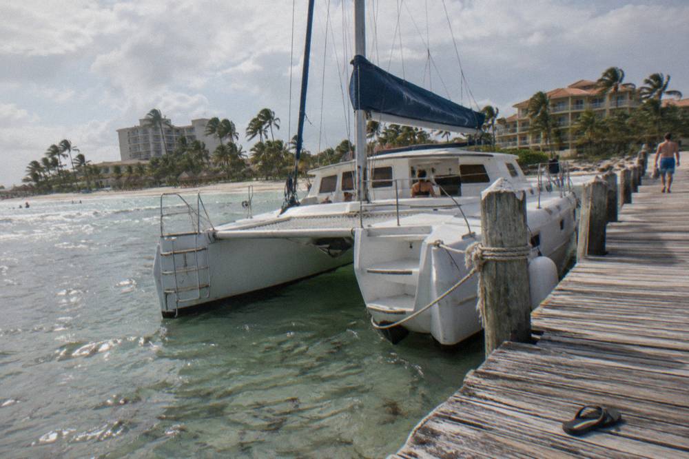 A catamaran moored at a wooden pier with calm shallow water and resort towers behind