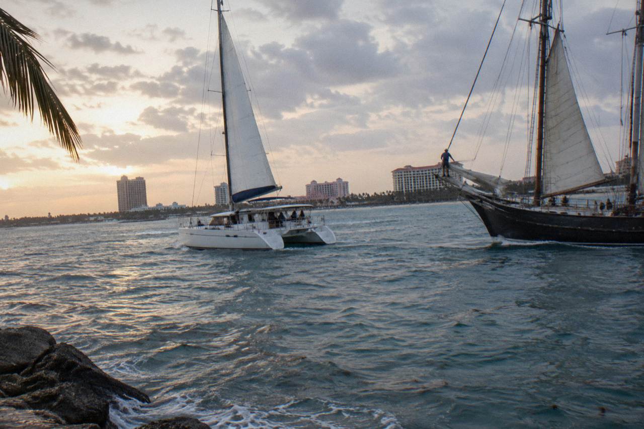 A catamaran and a schooner sail side by side on calm Caribbean water at dusk