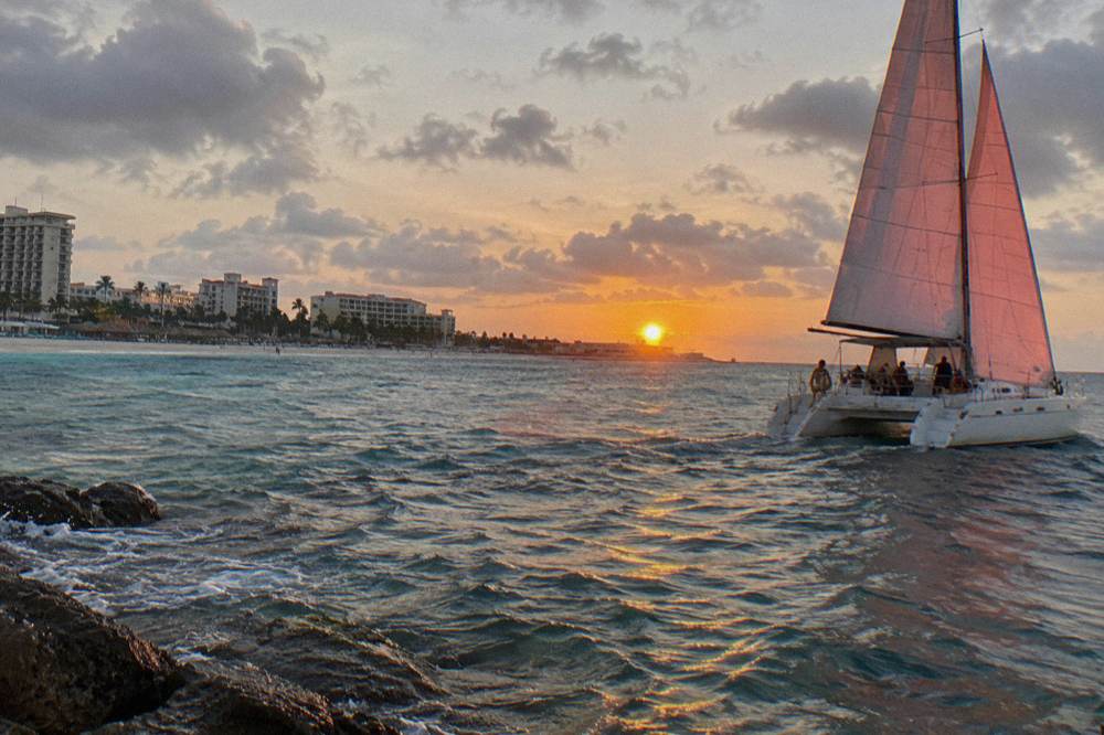 Catamaran under sail on open water with the sun nearing the horizon
