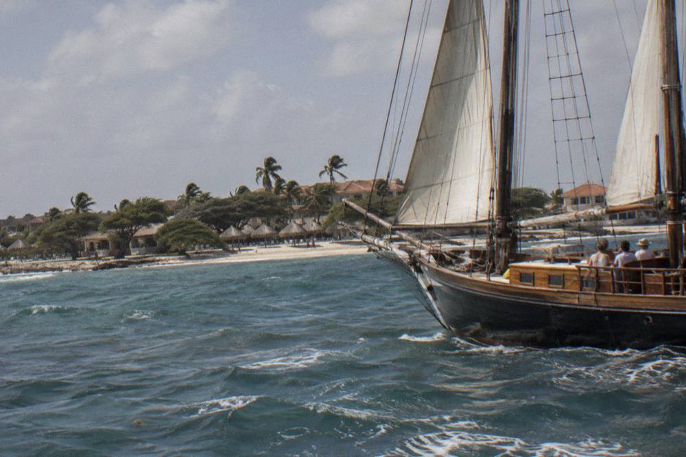 A wooden pirate schooner with tall masts and rigging sailing close to the Palm Beach coastline in late afternoon light