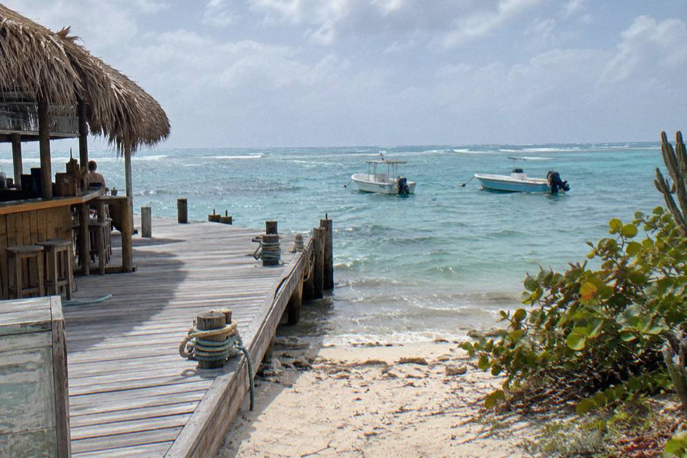 A thatched beach bar beside a wooden dock, with calm sea and two moored boats