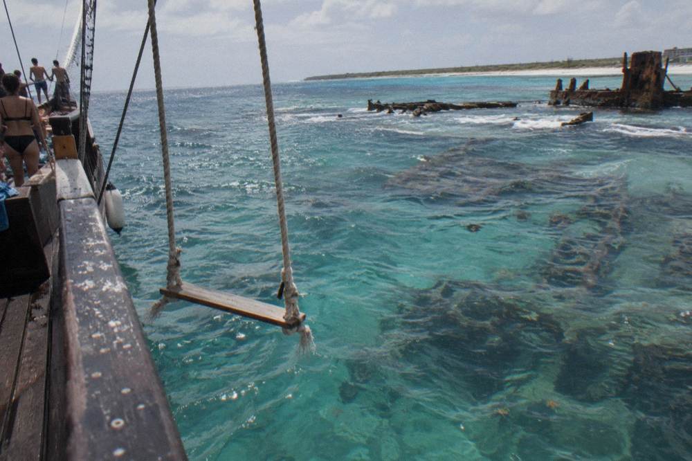 A rope swing extended over clear shallow water from the side of a wooden sailing ship with guests gathered on deck