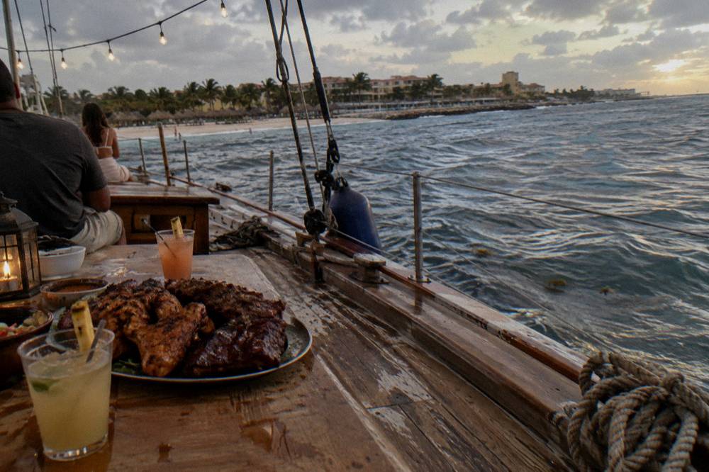 A buffet of grilled barbecue dishes and tropical cocktails arranged on the wooden deck of a sailing ship at dusk