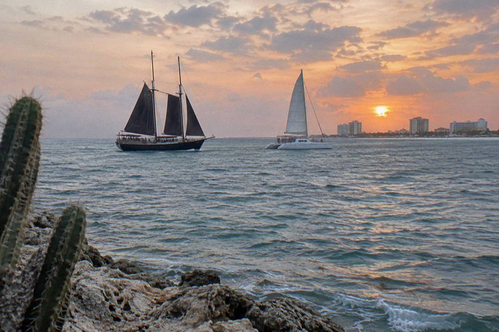 A pirate schooner and a catamaran sailing parallel along a coastline under a wide orange and pink evening sky