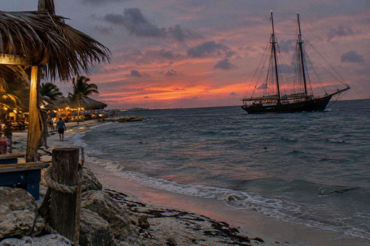 A wooden pirate-style schooner with tall masts anchored off a Palm Beach shoreline at dusk