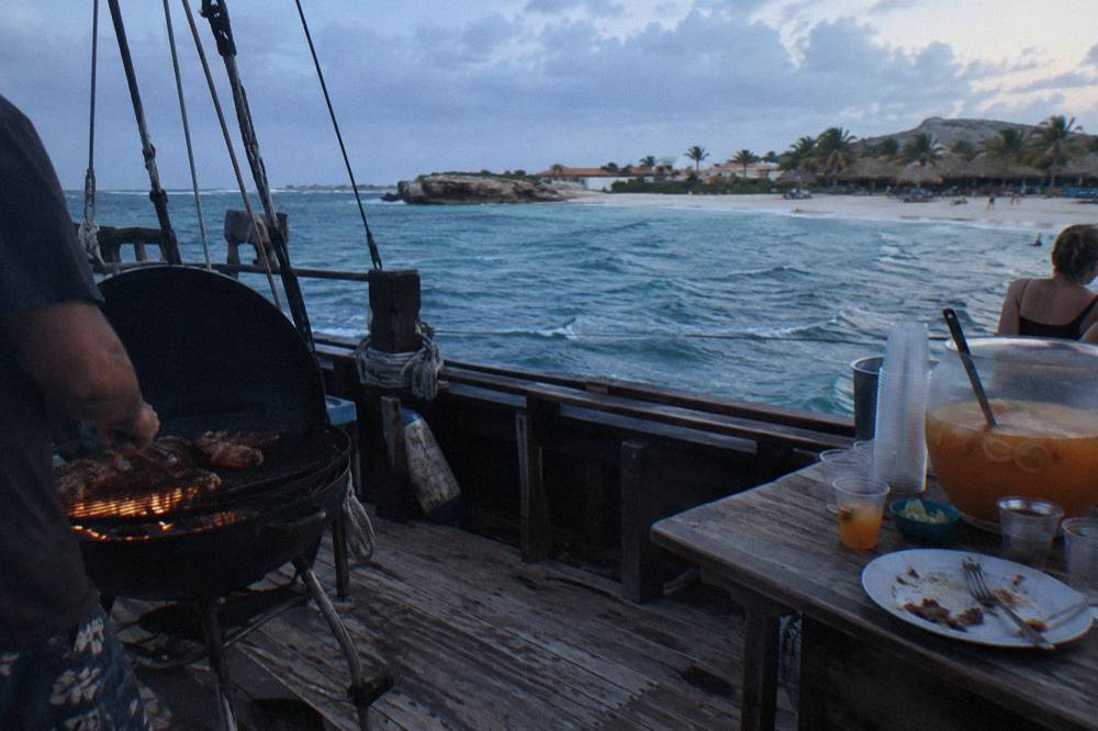 A lit barbecue grill and drinks table set up on a wooden ship deck at dusk