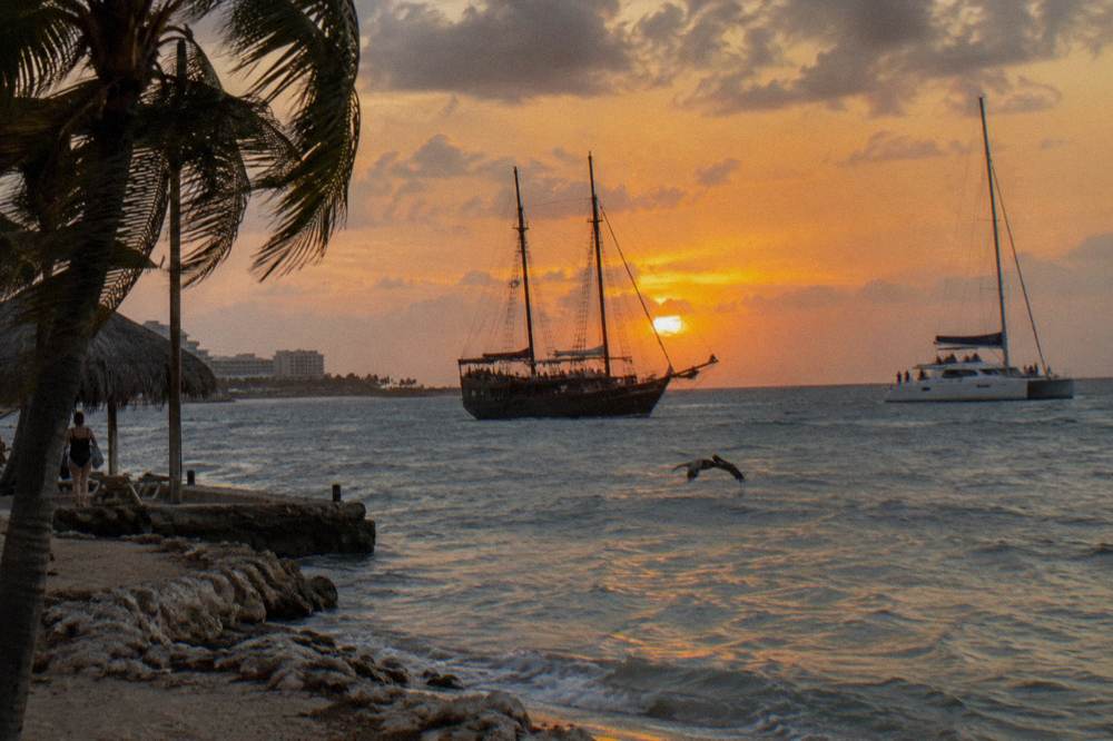 Two sailboats silhouetted against a bright orange sunset over calm Caribbean water