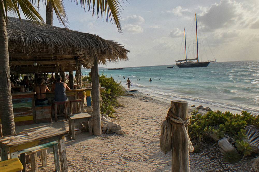 A sandy beach entrance with a thatched beach bar and a moored sailing ship in the background