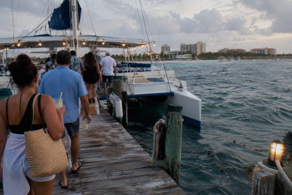 Open deck catamaran at a wooden pier with guests holding drinks before an evening sail
