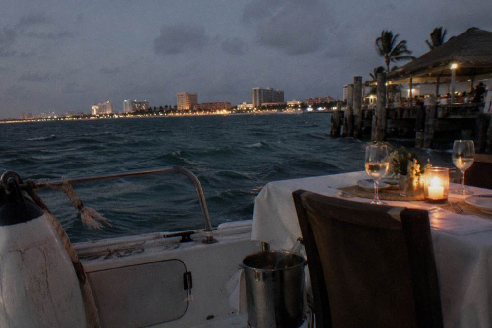 A dinner table set on a boat deck with glasses and candlelight beside dark water