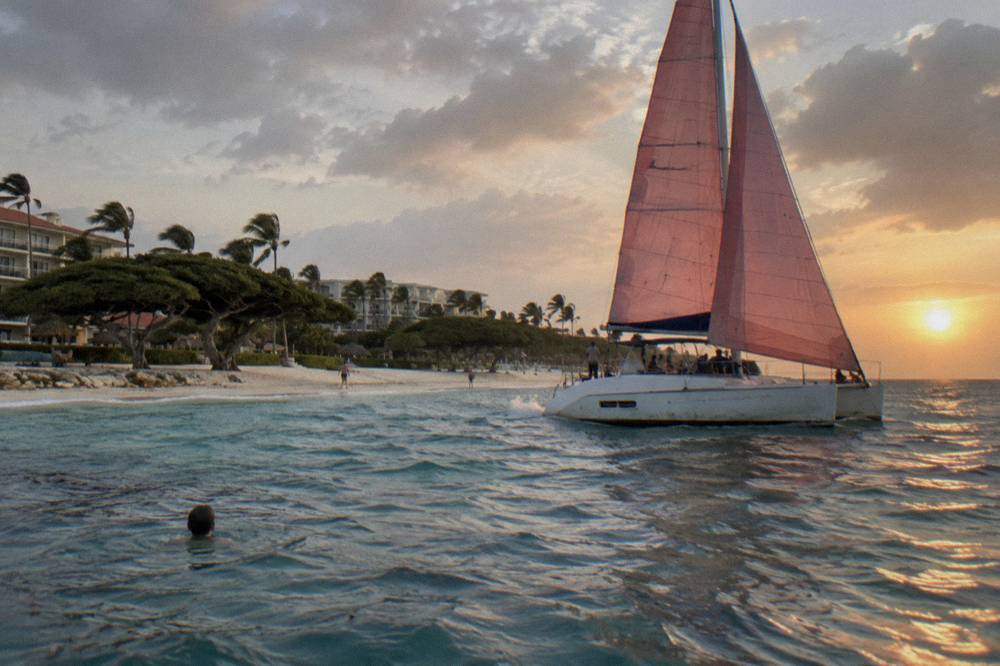 A catamaran under sail close to shore with calm water and a low sun over the horizon