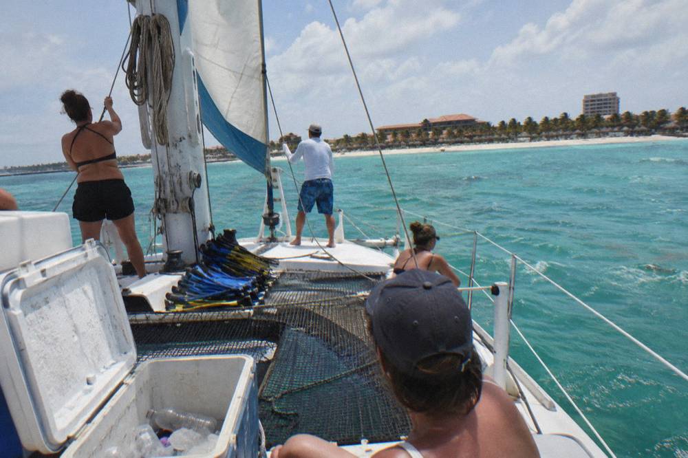 Passengers on a catamaran deck with sails raised cruising along a calm coastline in late afternoon light