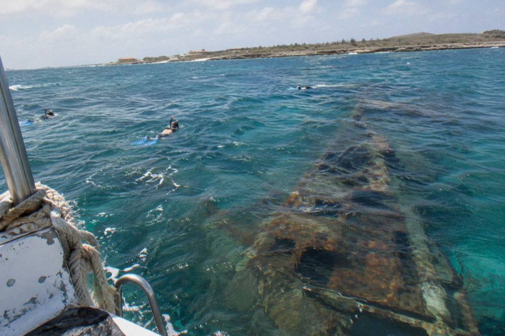 The dark hull of a sunken ship just below the surface of clear blue water