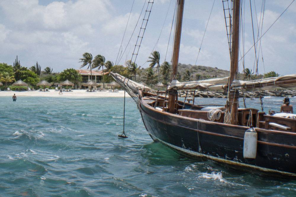A wooden schooner anchored near a Caribbean beach with palapas and swimmers