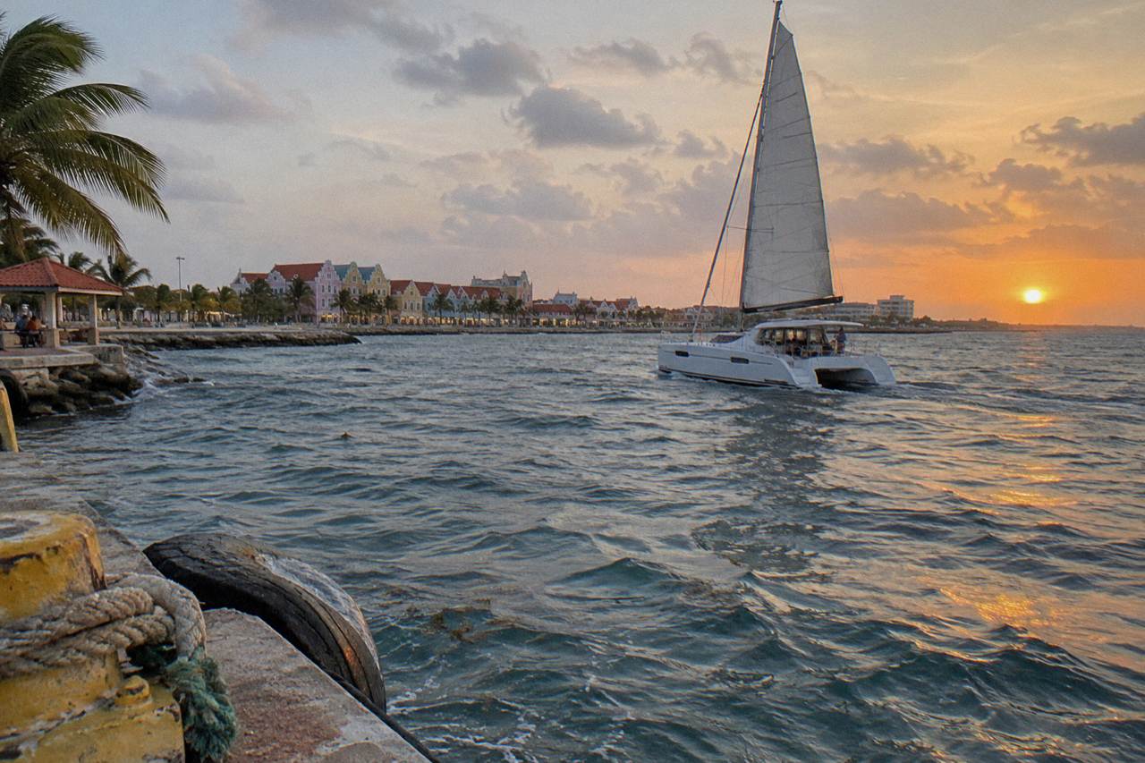 A catamaran under sail off the Aruban coast near dusk with calm water and a low sun