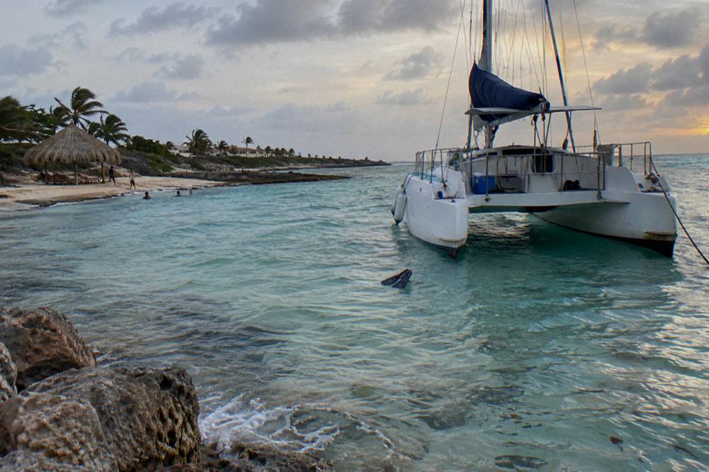 A white catamaran anchored near a beach with a thatched hut at dusk