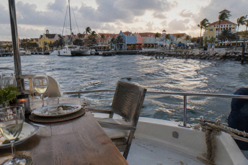 A dinner table set on a boat deck with plates and glasses as evening light fades over the sea