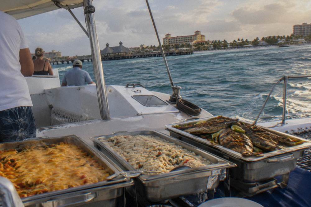 A buffet table with chafing dishes and plates of Caribbean food set out on a boat deck
