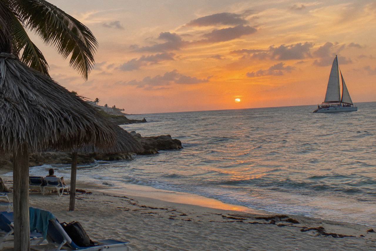 A catamaran under sail on calm water as the sky turns orange near the Aruba coastline