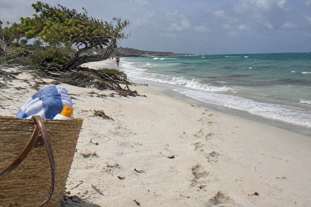 Beach tote bag with sunscreen bottle and rolled towel on white sand beside calm water