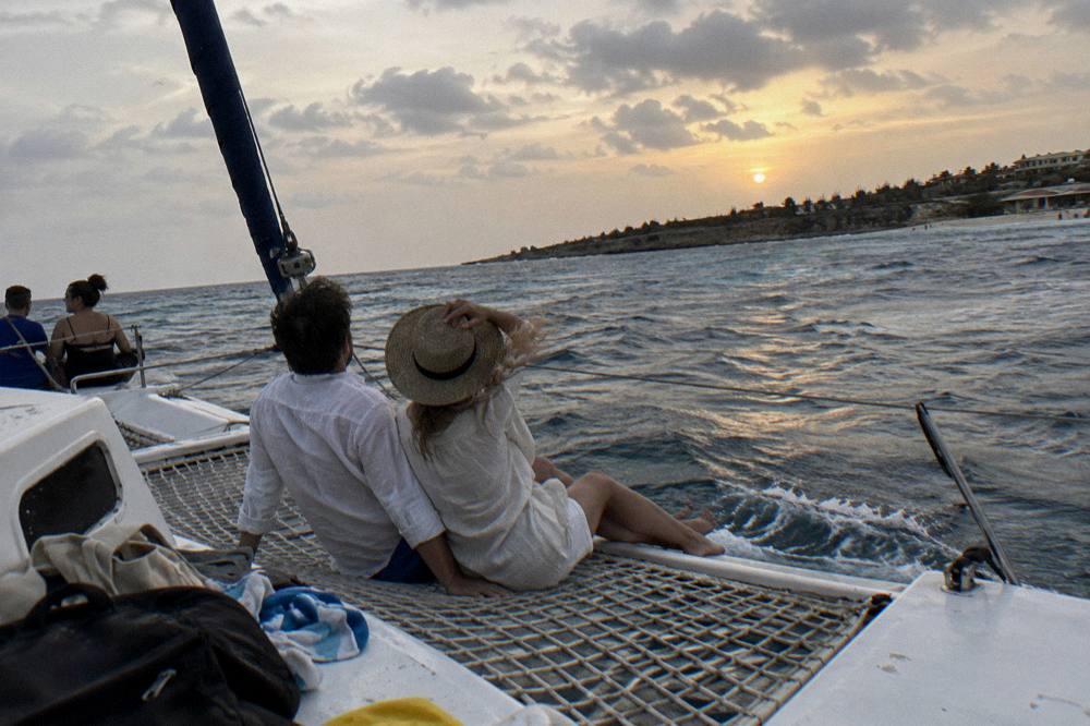 Couple in light summer clothing sitting on the bow of a boat facing an orange evening sky